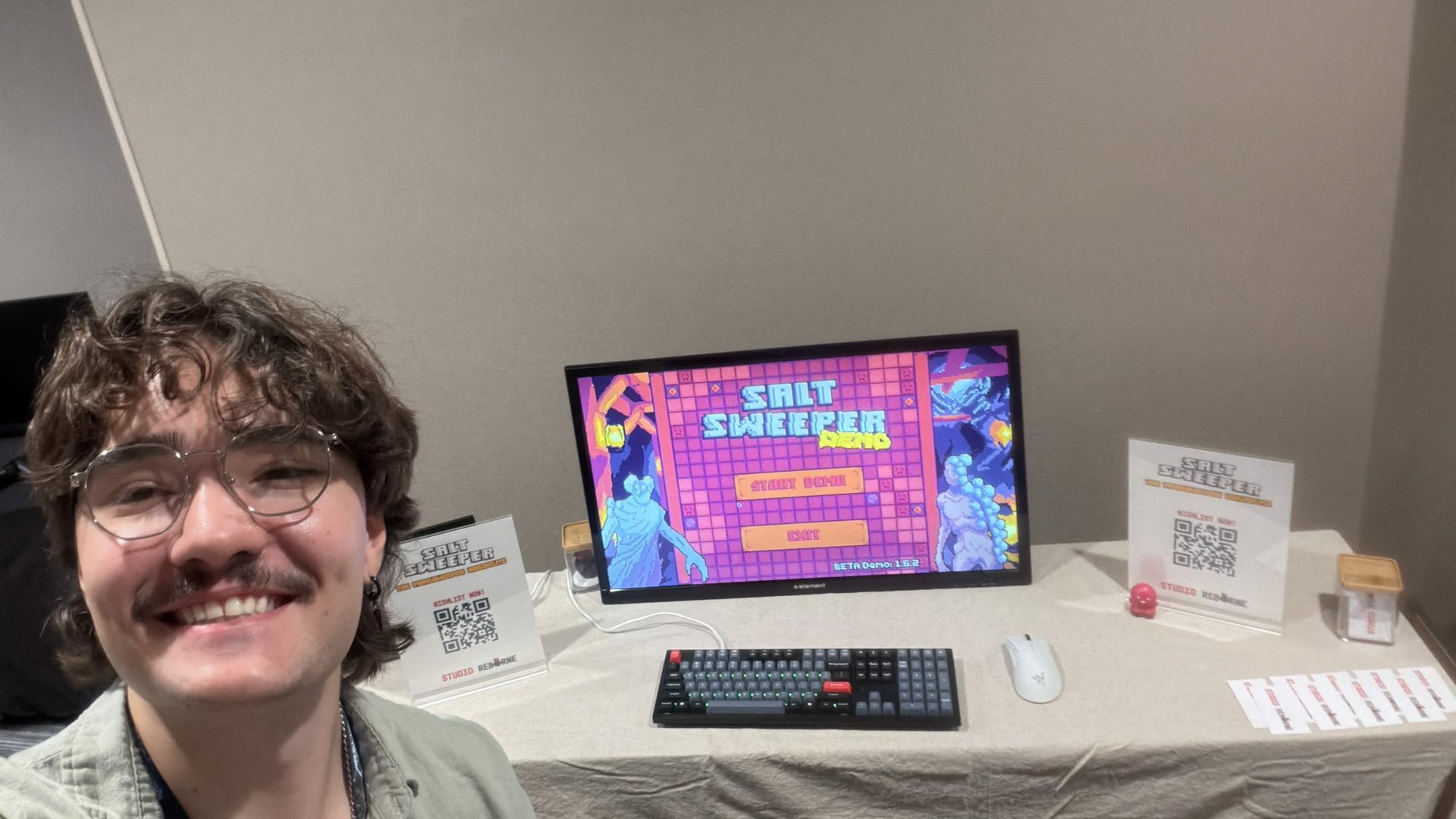 Corvin at the Salt Sweeper expo table, with the demo title screen, QR code signage, and business cards